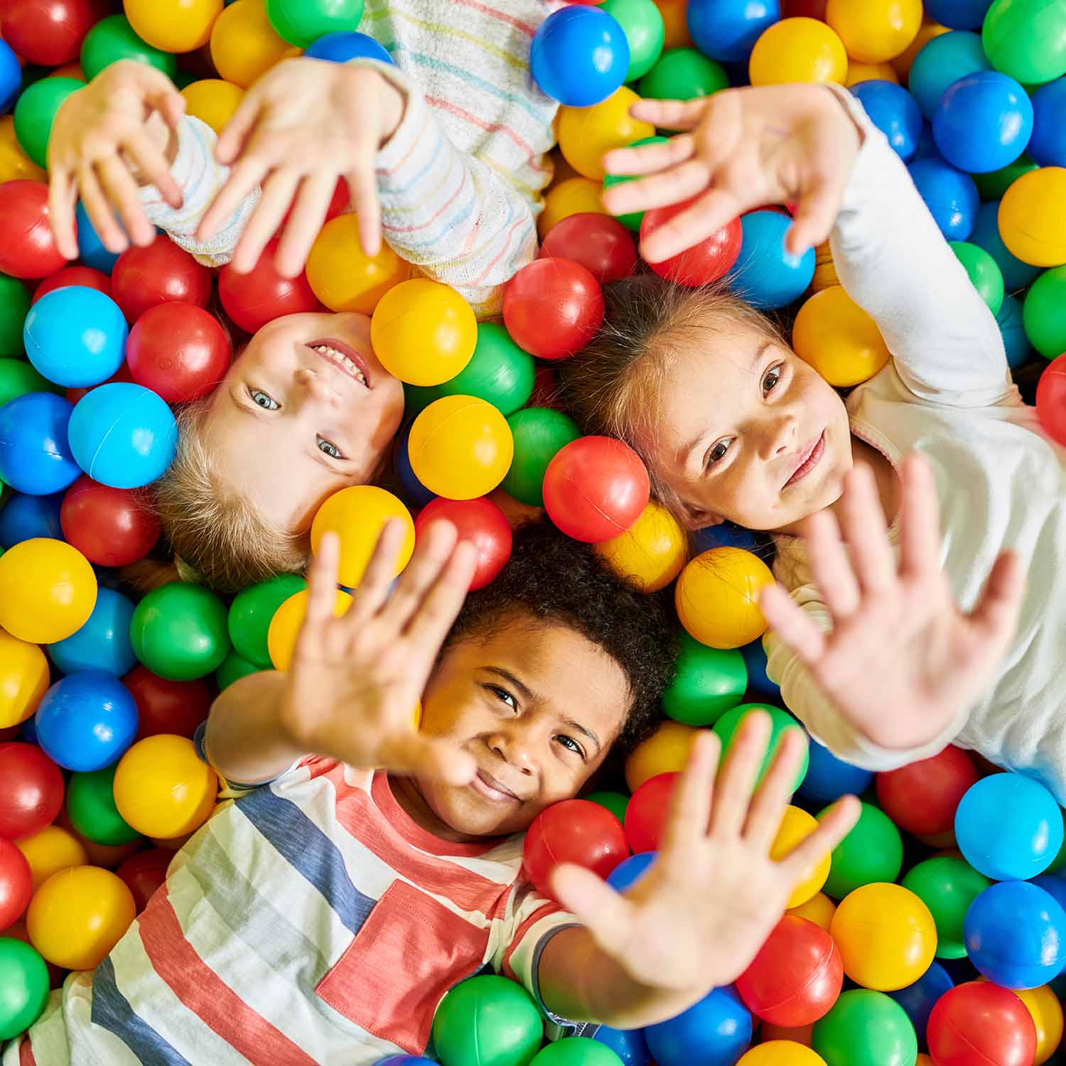 Drei Kinder spielen glücklich in einem bunten Bällebad und strecken die Hände in die Kamera.